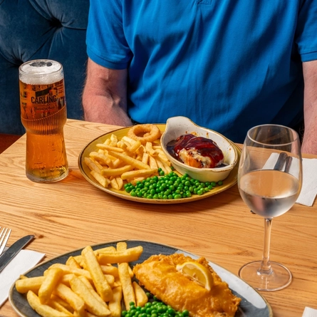 A plated portion of fish and chips and Hunters chicken with drinks in front of people on an indoor table.