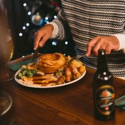 A group of people seated at a festive decorated table near a Christmas three. There are plates of food and drinks in front of them.