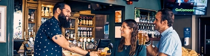 A member of staff serving two customers at a bar table.