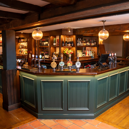 The wood panelled bar inside The Blue Cap, with wooden beams on the ceiling, and bottles of wine and alcoholic spirits on wooden shelves behind the counter.