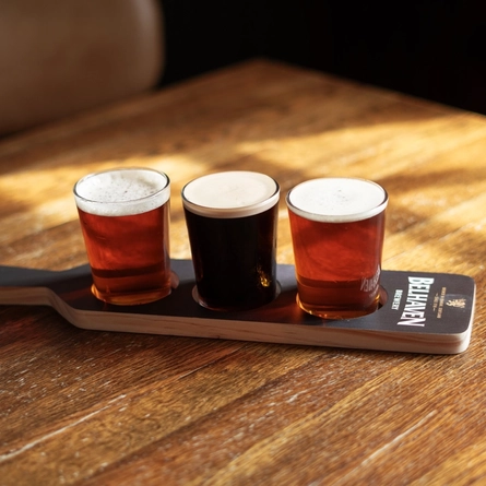 Three half pint glasses of ale or craft beers sitting in a paddle shaped drinks holder on a wooden table inside a pub.