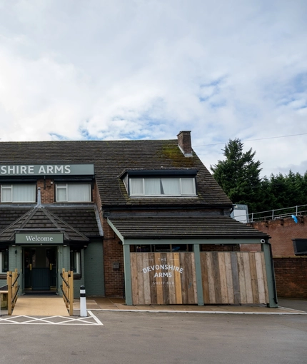 The exterior facade, signage, and seating area at the Devonshire Arms in Sheffield, with an accessibility ramp, wooden picnic tables, shade umbrellas, and string lights.