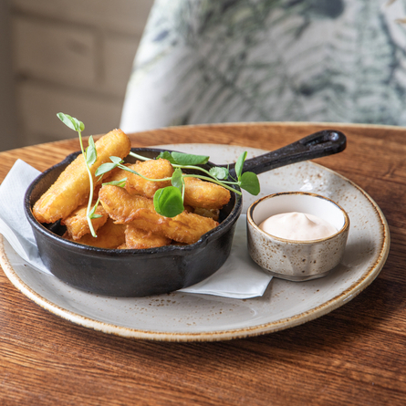 Halloumi fries served in a small skillet. The skillet sits on a plate with a small ramekin of dipping sauce and the plate sits on a wooden table.