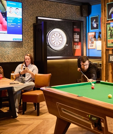 The interior pub gaming area at The Carousel, with a pool table, dartboard, TV on the wall, and restaurant tables. Two people are playing pool and other people are sitting at some of the tables.