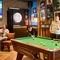The interior pub gaming area at The Carousel, with a pool table, dartboard, TV on the wall, and restaurant tables. Two people are playing pool and other people are sitting at some of the tables.