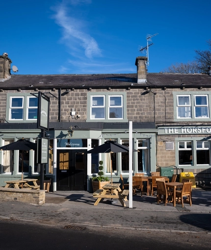 The exterior facade, signage, and seating area of The Horsforth, with wooden picnic tables, shade umbrellas, and strings lights above the seating area.