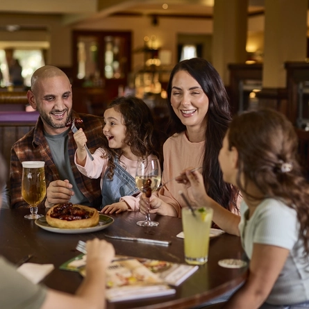 Three adults and three children sitting around a wooden restaurant table inside a Farmhouse Inns venue. Glasses of beer, white wine, and a soft drink sit on the table; as well as a plate of pigs in blankets served in a giant Yorkshire pudding.