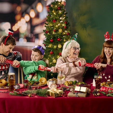 Three adults and a child sitting side by side at a restaurant table and pulling Christmas crackers together. The table is laden with plates of Christmas dinner; glasses of wine, beer, fruit juice, and soft drink; pine cones; and golden Christmas presents; and a Christmas tree is visible in the background.