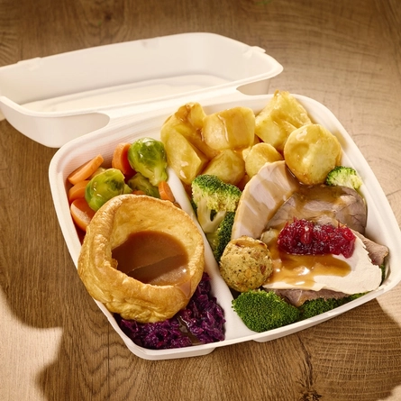 A close up view of a takeaway box filled with Yorkshire pudding, roast potatoes, broccoli, carrots, Brussels sprouts, stuffing, slices of roast meat, cranberry sauce, and gravy; sitting on a wooden table.