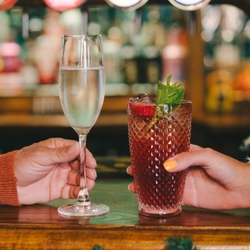 A glass of champagne and a cocktail served in a highball glass and garnished with mint, sitting on a bar inside a pub. A hand holds each glass and a Christmas garland is attached to the bar.