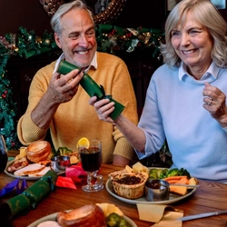 An image focusing on 2 people sat with others at a table within the interior restaurant seating area pulling a cracker while enjoying drinks and food dishes from the Festive Fayre menu at a Community Social venue.