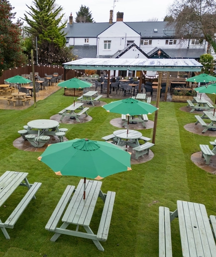 An aerial view of the exterior beer garden seating area at the Tredegar Arms, including the picnic tables and shade umbrellas on the lawn, the paved seating area, and the undercover area.