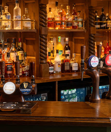 A closed up view of the wood panelled bar inside Ye Olde Swan in Woughton-On-The-Green, with bottles of wine and alcoholic spirits on wooden shelves behind the counter.