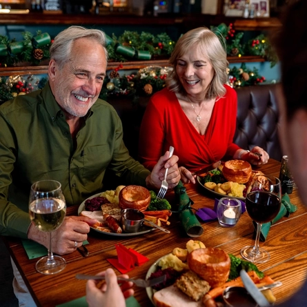An image focusing 2 people sat within a group at a table within the interior restaurant seating area enjoying drinks and dishes from the Christmas menu at a Community Social venue.