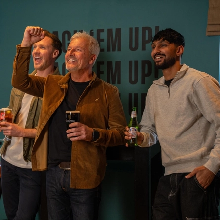 Three people standing in the gaming area of a restaurant. One of them is holding a glass of beer, one is holding a glass of Guinness, and the other is holding a bottle of Peroni.