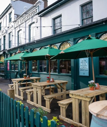 A close up view of the wooden picnic tables with shade umbrellas, outside in front of The Grosvenor. A potted plant sits on each table, and there are string lights overhead.