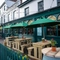 A close up view of the wooden picnic tables with shade umbrellas, outside in front of The Grosvenor. A potted plant sits on each table, and there are string lights overhead.