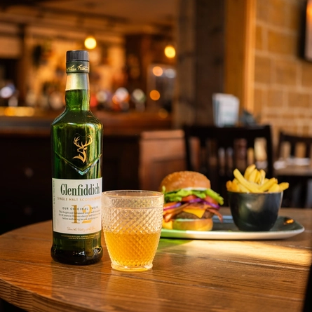 A served glass of Glenfiddich with the bottle next to it in the interior restaurant and seating area of a Community Pubs with Food venue, a blurred burger meal sits in the background.