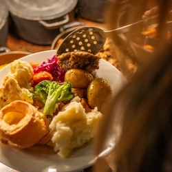 An over the shoulder view of a person standing at a carvery counter, holding a plate of food in one hand and using a large spoon to put stuffing onto the plate.