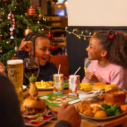 Two children sit at a festive dining table beside a decorated Christmas tree adorned with red and gold baubles, candy cane ornaments and warm fairy lights. One child holds up a breaded snack while the other holds a chip. The table is set with burgers, chips, vegetables, gravy, fruit juice cartons, wine and beer. Christmas-themed activity sheets and seasonal decorations are visible on the table, creating a warm restaurant setting decorated for the festive season.