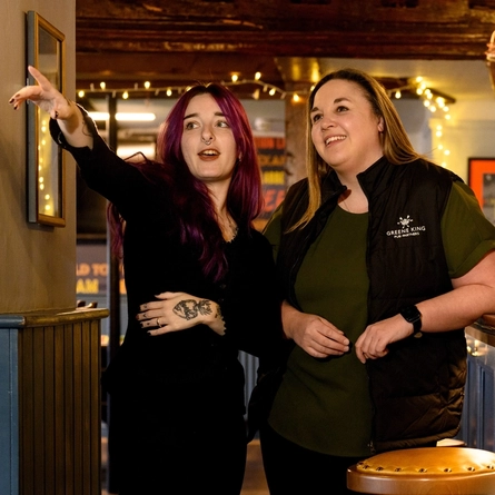 Two Women stood by the bar, the woman on the left is pointing with her arm extended. 