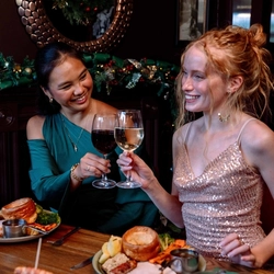 An image focusing on 2 people sat at a table with others holding glasses of wine, celebrating with a cheers while enjoying New Years Eve dishes within the interior restaurant seating area at a Community Social venue.