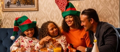 A festive restaurant booth decorated for Christmas, with four people seated around a wooden table. Two people wear green-and-red elf hats, while another wears Christmas-themed pyjamas. Plates of breakfast food, a glass of orange juice, a juice carton, Christmas crackers, colouring materials and a small reindeer toy are arranged on the table. A red Christmas stocking hangs on the patterned wall behind the group, with framed artwork and string lights completing the seasonal setting.