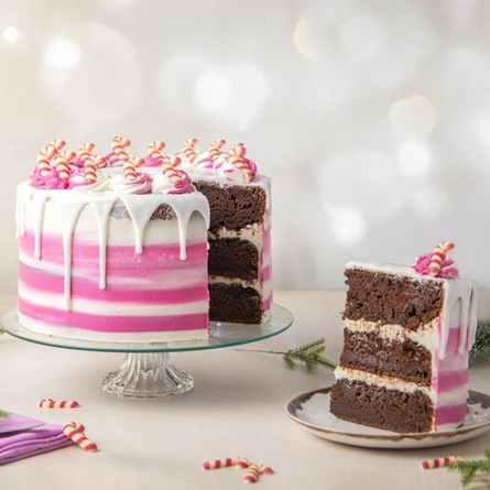 The Candy Cane Lane cake served on a glass cake stand which sits on a table scattered with pine tree sprigs and mini candy cane decorations. A slice has been cut out of the cake and served on a plate sitting on the table next to the cake stand.