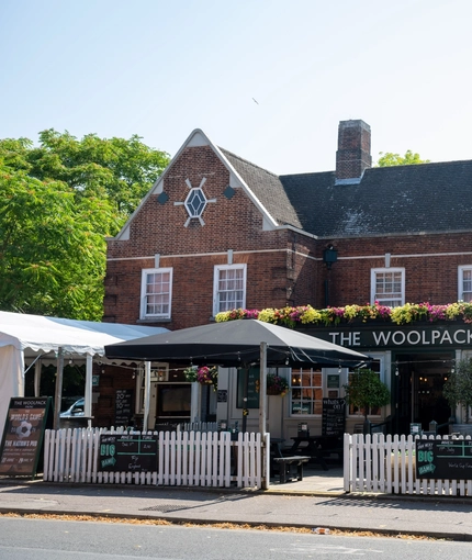 The exterior facade, signage, and beer garden seating area of The Woolpack Inn in Norwich; with trees on each side of the entrance, string lights above the tables and chairs, and flowers baskets on the walls.