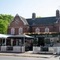 The exterior facade, signage, and beer garden seating area of The Woolpack Inn in Norwich; with trees on each side of the entrance, string lights above the tables and chairs, and flowers baskets on the walls.