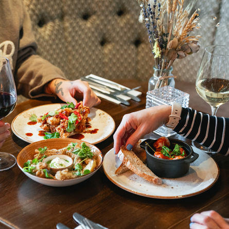 Three meals served in plates and a bowl, sitting on a restaurant table next to cutlery, table decorations, and glasses of red and white wine. Two people are sitting at the table and picking up food from the plates in front of them.