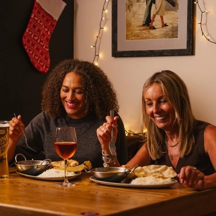 Two diners seated at a wooden table enjoy a festive curry meal in a warmly decorated restaurant. The table is set with metal curry bowls, plates of white rice, naan bread, a glass of rosé wine and a pint of beer. Christmas decorations include a red stocking hanging on the wall, warm string lights, and a small tabletop Christmas tree with candy cane ornaments. A framed artwork hangs above the table, adding to the cosy festive setting.