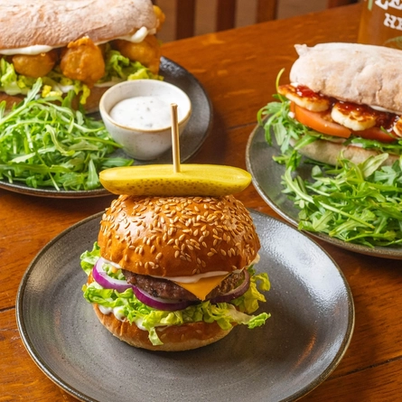A Cheeseburger, a Cod Goujon Ciabatta, and a Halloumi Ciabatta, each served on a plate which sits on a wooden restaurant table along with a glass of Level Head.