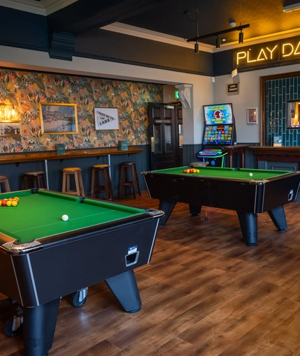 The interior games area at The Victoria, with two pool tables, a gaming machine, bar stool seating, and a neon sign which reads "Play, Darts, Pool".
