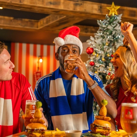 Three football fans sit around a wooden table in a sports pub decorated for Christmas, with a frosted Christmas tree and live football shown on large screens in the background. One person wears a Santa hat, another wears reindeer antlers, and team scarves add to the matchday atmosphere. Burgers topped with onion rings and pickle skewers, chips and glasses of beer are served on the table as the group celebrates during a festive Boxing Day football gathering.
