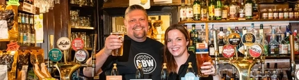 A man and woman holding pints of beer behind a bar.