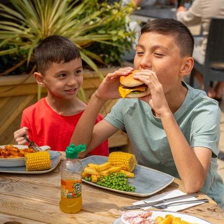 A boy seated at an outdoor table in a beer garden, eating a burger whilst his younger brother looks on..
