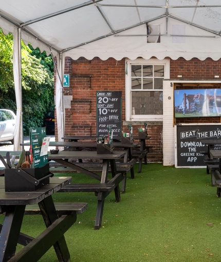 The exterior beer garden seating area under a large gazebo, at The Woolpack Inn in Norwich; with wooden picnic tables, and a TV on the wall.