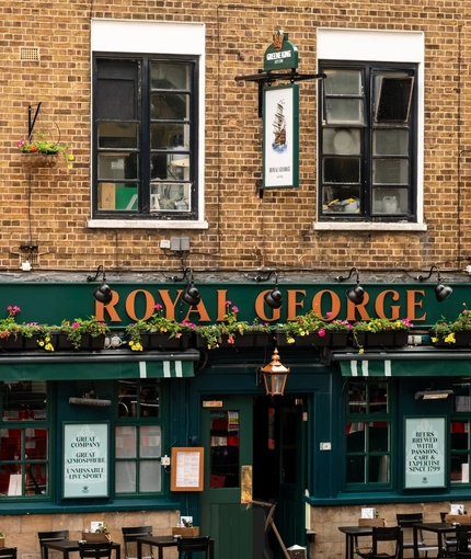 The exterior facade, signage, and seating area of the Royal George in Euston; with a large golden lantern hanging above the door, and flower boxes above the windows.