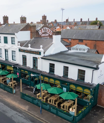 An aerial view of the exterior facade, signage, and seating area of The Grosvenor, with wooden picnic tables in front of the pub, shade umbrellas, and string lights above the seating area.