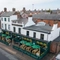 An aerial view of the exterior facade, signage, and seating area of The Grosvenor, with wooden picnic tables in front of the pub, shade umbrellas, and string lights above the seating area.