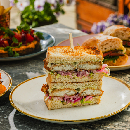 An advertising image showing three different sandwich dishes including Heritage Tomato Sandwich, Sirloin Steak Ciabatta, Panko Haddock & Ham & Emmenthal Croque Monsieur sat on a table within the exterior beer garden seating area at a Crafted venue.