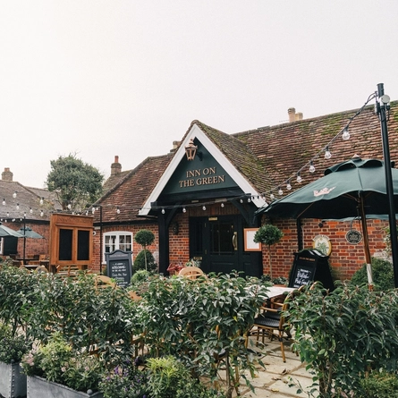 The exterior facade, signage, and beer garden seating area of The Inn On The Green in Harpenden, with wooden furniture, shade umbrellas, shrubs in planter boxes, and string lights overhead.