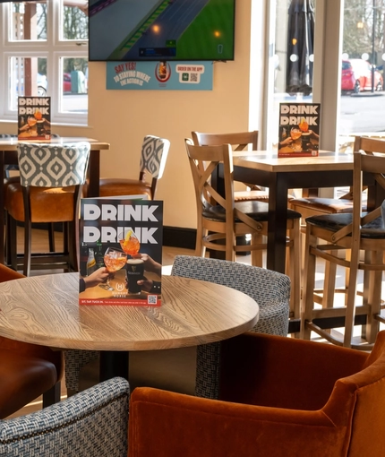 An interior restaurant seating area at The Weathervane, with upholstered chairs, a dartboard, and a TV on the wall.