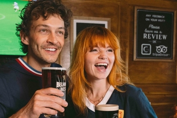 An image focusing on 2 people within a group of friends stood at a table watching a football match and enjoying a selection of drinks and snacks within the interior seating area at a Greene King venue.