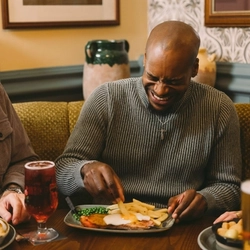 Three people dining together at a pub table, with plated meals, chips, peas and glasses of beer.