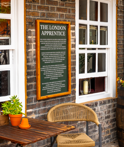 A close up view of one of the wooden tables and wicker-style chairs next to the exterior brick wall of The London Apprentice. Menus and a potted plant sit on the table; a sign on the wall details the history of the pub; and a large beer barrel filled with plants and flowers stands nearby.