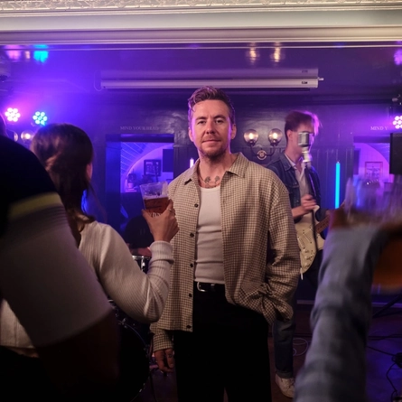 A live music performance in a pub venue, with Danny Jones standing among audience members in front of a small stage, smiling at the camera. A guitarist, drum kit and microphone stand are visible in the background, while purple and blue stage lighting illuminates the room. Audience members holding drinks surround the performance area, creating an intimate live entertainment atmosphere.
