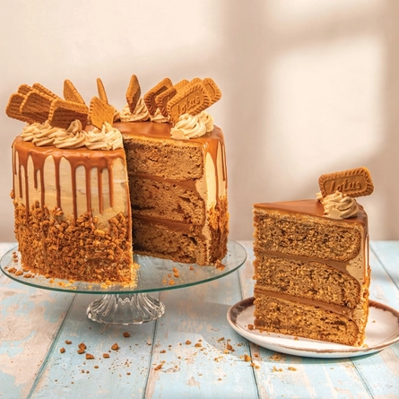 A Biscoff cake on a glass cake stand, on a table next to a slice of the cake served on a plate.