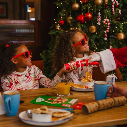 A festive breakfast table set beside a decorated Christmas tree adorned with red and gold baubles, candy cane ornaments and warm fairy lights. Two children wearing Christmas-print pyjamas and red novelty sunglasses sit at the table, with one holding a red Christmas cracker being handed over by a person dressed as Santa Claus. Breakfast dishes, mugs, juice cartons, a glass of orange juice, Christmas crackers and a colouring activity sheet are arranged on the wooden table, while other diners sit nearby in the Christmas-themed restaurant setting.